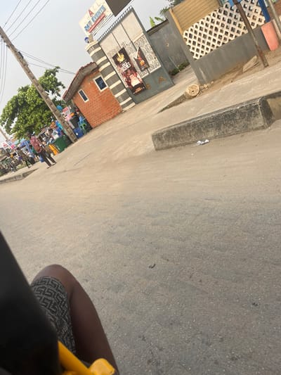 The image displays a street scene in Isasi, Nigeria, under a hazy sky. In the lower left foreground, a dark-skinned leg in patterned shorts and a yellow vehicle component are visible, indicating the perspective is from inside a vehicle. The midground consists of a grey asphalt road, with a long, grey concrete curb situated on its right edge. Beyond the road, to the left, a wooden utility pole supports multiple wires next to green trees. Adjacent to the trees, a cluster of market stalls is present, displaying various items and occupied by several individuals; one person in a patterned shirt is observed walking towards the right. Centered in the background is a small, red-brick building with a red-tiled roof and a single window, featuring grey striped accents. This structure abuts a large, ornate grey metal gate, which displays two vertical posters, one clearly advertising "247 EVENTS" and another depicting "SCENES". Above the gate, a larger sign is mounted, also reading "247 EVENTS RENTALS & TRAINING CENTER". To the far right of the scene, a grey wall with a white lattice pattern on its upper section is visible.