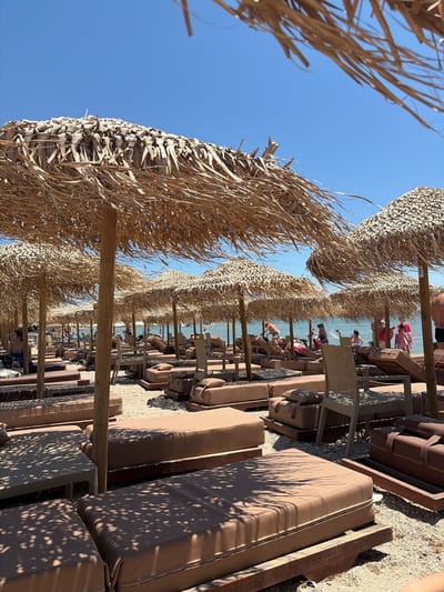 The image depicts a beach scene in Artemida, Greece, under a clear blue sky, suggesting a sunny day, likely late morning or early afternoon given the bright light and shadows. The foreground is dominated by rows of thatched umbrellas providing shade over empty lounge chairs. These chairs, upholstered in a muted brown fabric, are arranged in parallel lines, facing a body of water visible in the background.
Beyond the lounge chairs, people are present, scattered along the sandy shore and in the shallow water. Some individuals are seated on the chairs, while others are standing or moving about. A few people are visibly interacting with the water, with one person appearing to toss something. The overall atmosphere suggests a relaxed and leisurely environment, typical of a beach resort.
The thatched umbrellas are constructed from dried palm leaves or similar natural materials, casting dappled shadows on the sand and chairs. Wooden poles support these umbrellas, creating a structured, organized layout. The sand itself appears to be a mixture of fine grains and small pebbles.
In the distance, the sea is calm and appears to stretch out towards the horizon. The presence of lounge chairs and umbrellas indicates this is likely a managed beach area, possibly part of a hotel or a beach club. No visible text is present in the image. The overall impression is one of a serene summer day at a popular Mediterranean beach.