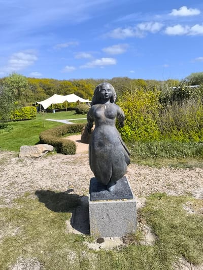 The image shows a bronze statue of a woman standing on a gray concrete pedestal. The statue is located in a garden setting near Sint Maartensvlotbrug, Netherlands. Behind the statue is a green lawn, a winding path bordered by shrubs, and a white tent-like structure. Trees and bushes surround the area. The sky is blue with some clouds.