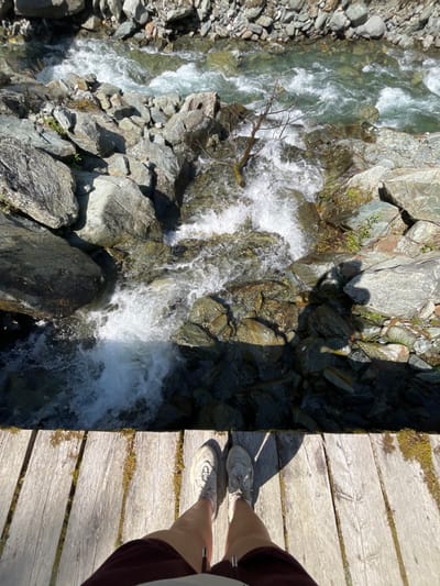 The image captures a downward-facing perspective from a wooden bridge over a rocky, fast-flowing river in Reitlehen, Austria. In the foreground, the lower legs and feet of an individual are visible, standing on weathered wooden planks. The person is wearing light-colored athletic shoes and dark-colored shorts. Their shadow is cast forward and to the right on the wooden surface. Below the bridge, a clear, turbulent river cascades over numerous grey and dark grey rocks, creating white rapids and foamy sections. A single, bare, broken tree branch or small trunk is partially submerged and entangled among the rocks in the central part of the river. The environment is well-lit by direct sunlight, casting distinct shadows and highlighting the texture of the rocks and the agitation of the water.