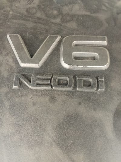 This media file shows a close-up of raised lettering on a dark, textured surface, likely a component of a vehicle engine. The text "V6" is prominently displayed in large, silver-colored capital letters. Below "V6," the word "NEODi" is also visible in smaller, silver-colored capital letters. The lighting suggests it is daytime, and there are no obvious cues about the weather. The setting appears to be an automotive context, possibly under the hood of a vehicle. There are no people visible in the media file. The surface is dusty and shows some wear, which is typical for engine components. The overall impression is one of identification of a specific engine type or technology. The provided location context of Alatau, Kazakhstan, does not offer any further visual clues within the image itself.