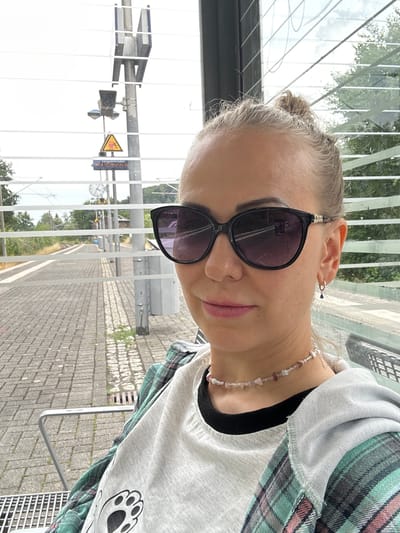 In Velbert, Germany, a young woman with blonde hair tied in a bun sits at a train station platform, evidenced by the tracks and signage. She is wearing large, dark-rimmed sunglasses, a gray t-shirt with a paw print graphic, and a teal and pink plaid shirt over it. A delicate beaded necklace adorns her neck, and a small hoop earring with a dark blue charm is visible in her left ear. The woman is positioned in the foreground, with the train tracks and platform extending into the background. The scene is set within an outdoor shelter, indicated by the slatted glass panels and metal seating. A traffic sign with a yellow triangle and a red symbol is visible on a pole in the background, along with a clock and railway signals. The ground is paved with gray bricks.