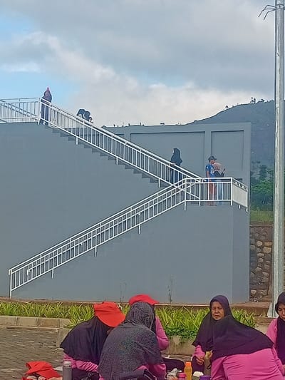 The image depicts an outdoor scene in Pangalengan, Indonesia, featuring a group of people engaged in various activities near a modern grey concrete structure with prominent white metal stairs and railings.
In the foreground, a group of approximately five women is seated on a paved surface, seemingly enjoying a picnic or gathering. Most are wearing hijabs; one woman wears a bright red hat, another a black patterned hoodie, and a woman in a purple hijab looks towards the left with a neutral expression. Several bottles and containers are visible on the ground near them, suggesting food and drink. Behind this group, there is a neatly maintained strip of green foliage.
On the grey concrete structure, other individuals are observed. At an intermediate landing, a man in a blue t-shirt and cap stands next to a woman in a beige hijab, both looking intently at a smartphone held by the woman. Further up the stairs from this landing, another person in a black hijab is ascending. Near the very top of the stairs, a solitary figure appears to be holding a phone, possibly taking a selfie or observing the surroundings.
The background reveals rolling hills or mountains under an overcast, cloudy sky. A utility pole stands on the far right side of the frame. The scene conveys a sense of leisure and social interaction in a scenic, natural environment, possibly a newly developed recreational area given the clean lines of the structure. No discernible text is visible in the image.