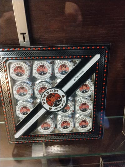 A box of "Imperador" chocolates from "Bom Bom Avianense" is displayed. Each chocolate is individually wrapped in silver foil with a circular label featuring a portrait and the brand name. The box is black with a decorative red border and a white and black diagonal band across the center. A "T" sign is visible above the box, which sits on a glass shelf against a wood-paneled background.