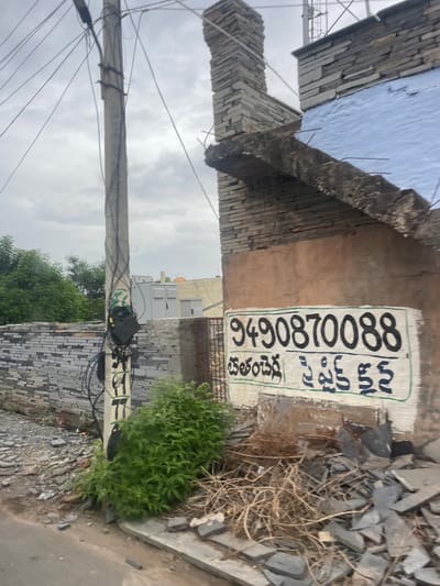 The image shows a roadside scene in Bethamcherla, India. A concrete utility pole stands on the left, covered in tangled electrical wires. To the right of the pole, a building with a light blue, textured roof and a brick chimney is visible under construction or repair. A portion of the building's wall is painted in a pale orange hue, and a white sign with a green border is affixed to it. The sign displays a phone number, "9490870088," in bold black characters at the top. Below the number, text is written in what appears to be Telugu script, with the words "Bethamcherla" and "Spick." The ground in the foreground is littered with debris, including broken grey tiles and dry branches, suggesting a construction site or an area undergoing renovation. Green foliage grows around the base of the utility pole and near the debris. The sky is overcast with clouds.