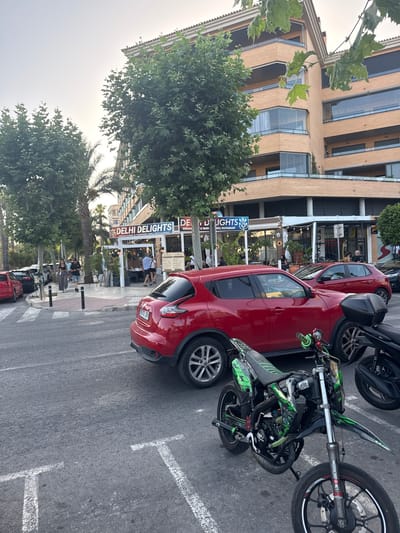 The image shows a street scene in l'Alfàs del Pi, Spain. In the foreground, a green and black dirt bike is parked on asphalt. Behind it, a red Nissan Juke is parked. Further back, another red car and a black motorcycle are visible. On the right side of the street, a multi-story building with a beige facade and balconies overlooks a row of shops. One of the shops is "DELHI DELIGHTS INDIAN CURRY & TANDOORI GRILL RESTAURANT". People are seen walking on the sidewalk and sitting at tables outside the restaurant. Trees line the sidewalk, and a palm tree is also present. The sky is overcast.