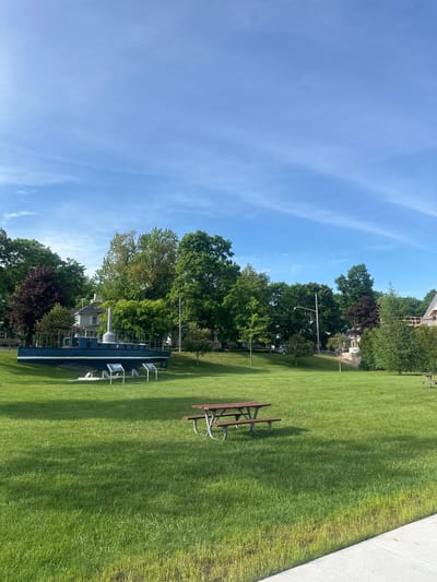 This is a photograph of the waterfront park in Harbor Springs, Michigan. The park features a preserved submarine, a picnic table, and a clear blue sky with wispy clouds. The submarine is dark blue with a white stripe and is mounted on a stand on a grassy hill. Two informational plaques are placed in front of the submarine. A wooden picnic table with benches sits on the lawn in the foreground. The background consists of lush green trees and houses. The sun is casting shadows on the grass.