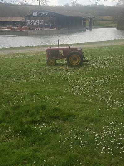 The scene depicts an outdoor environment in Avignonet-Lauragais, France, under natural, diffused light. In the foreground, a green grassy field is visible, densely interspersed with small white flowers. A stationary, weathered red tractor, featuring yellow wheel rims, is positioned in the mid-ground on this grass. A narrow dirt or gravel path separates the field from a body of water, identified as a pond or canal, which extends across the middle of the frame. On the opposite side of the water stands a dark-wood building with a brown, low-pitched roof, which appears to be an establishment with an open-air seating area. This area is furnished with multiple tables and chairs, and several individuals are observable, both seated and standing, on a concrete platform adjacent to the building. The building's facade displays several large, monochrome portraits or posters. In the background, a gently sloping, tree-covered hillside is visible under an overcast sky.