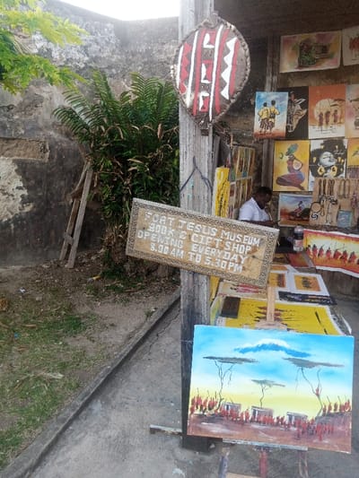 This image depicts an outdoor art and craft stall, confirmed by a visible wooden sign as the "FORT JESUS MUSEUM BOOK & GIFT SHOP," located in the Mji Wa Kale ward of Mombasa, Kenya.
One adult male is present, seated behind a display table. He has dark skin, short dark hair, and wears a white short-sleeved collared shirt. He is leaning forward, seemingly engaged with an item on the table.
The stall features numerous art pieces and crafts. A weathered wooden sign, framed and angled, hangs prominently, displaying the shop's name and operating hours: "OPENING EVERYDAY 8.00 AM TO 5.30 PM." An oval, shield-shaped decorative object with red, white, and brown geometric patterns hangs from a wooden post. Various unframed paintings, depicting African-themed subjects such as tribal figures, wildlife, landscapes with acacia trees, and stylized portraits, are displayed on a background wall and laid out on the table. A freestanding wooden easel showcases a landscape painting featuring a snow-capped mountain (likely Kilimanjaro), acacia trees, traditional huts, and numerous red-clad figures. A rack displays an assortment of necklaces and small craft items. A plastic water bottle is also visible on the table.
The background includes a tall, dark, weathered stone or concrete wall, partially obscured by dense green foliage, including a large bush. A small wooden ladder leans against this wall and vegetation on the left. The foreground consists of a concrete or paved surface, transitioning to dirt and patchy grass on the left. The scene is illuminated by natural daylight.