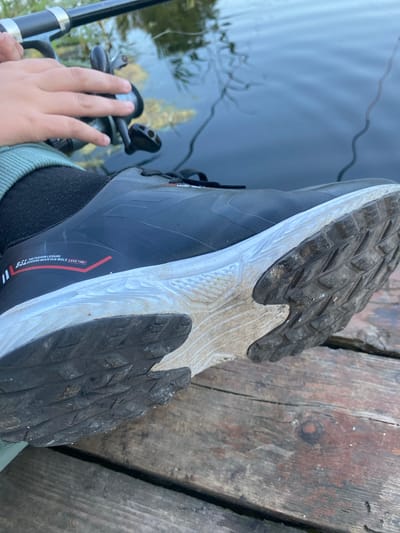 The media file shows a close-up view of a person's foot wearing a black waterproof boot and a dark sock, resting on a weathered wooden pier. The boot's sole, heavily textured for grip, is visible, showing signs of dirt and wear. To the left of the boot, a fishing rod with a reel is held by a hand, suggesting the person is engaged in fishing. The water is visible in the background, appearing calm with reflections of the surrounding environment. The scene is likely outdoors, given the natural light and wooden pier setting. The time of day appears to be daytime, with the sunlight creating soft shadows. The overall mood is relaxed, indicative of a leisurely activity like fishing. There is text on the side of the boot that reads "BSL the FASHION LEISURE MOUNTAIN WHOLE LEAD TIME". The location context is provided as Афанасьево, Russia.