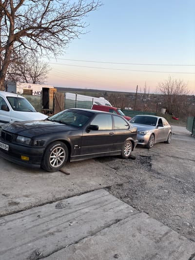 The image depicts an outdoor scene in Septemvritsi, Bulgaria, during twilight. The sky is light blue with a faint orange-pink hue near the horizon. The ground in the foreground consists of cracked concrete slabs, transitioning to an uneven surface of gravel, dirt, and potholes further into the mid-ground.

Three vehicles are prominently positioned: In the mid-ground, a dark-colored (likely black or very dark grey) BMW E36 Compact, bearing Bulgarian license plate 'P 5409 BH,' is parked on the concrete. A metal bar is placed underneath its front left tire, which displays 'HANKOOK' lettering on the sidewall. Immediately behind this vehicle, a silver BMW E60 sedan is parked on the gravel surface, facing the same direction. A white utility van is partially visible on the far left, parked behind the dark BMW.

In the background, a rustic, square-shaped structure made of concrete blocks and wood, featuring an open dark doorway and an 'HD' logo, is located on the left. A green metal mesh fence with concrete posts extends across the mid-ground, separating the vehicle area from a slightly elevated, sparsely vegetated landscape with bare deciduous trees and distant structures. A red pickup truck and another white van are partially visible behind the fence and the main vehicles. No individuals are present, and no active actions or interactions are depicted within the scene, which primarily shows stationary vehicles and their immediate environment.