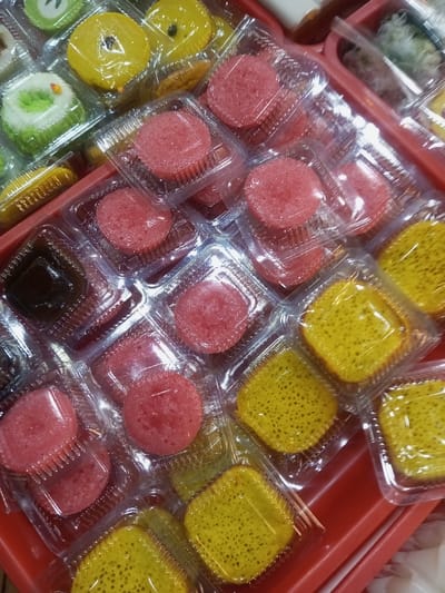 The image displays a dense arrangement of assorted Vietnamese desserts, individually sealed in clear plastic clamshell containers, situated on a red plastic tray. The majority of the frame is occupied by approximately four dozen packaged items. Dominant types include numerous round, pink/red spongy-textured cakes and multiple square, bright yellow cakes exhibiting a porous, honeycomb-like surface. Other visible items, primarily in the upper and lower left, include several round yellow cakes, square dark brown jelly-like confections, elongated yellow items, and a few green and white layered cakes with a central dark dot. The plastic containers feature uniformly ridged lower portions. The scene depicts a commercial food display within Lê Thanh Nghị Ward, Vietnam. No individuals or active interactions are visible.