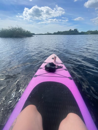 A first-person perspective shot shows a person on a pink paddleboard on a body of water, likely a lake or river, with trees and clouds in the background. The paddleboard has a black bag and a can on it. The person's legs are visible, with tattoos on their left leg. The water is choppy and reflects the sky. The image is likely taken in the city of Ozerki, Russia, given the surrounding vegetation and open water.