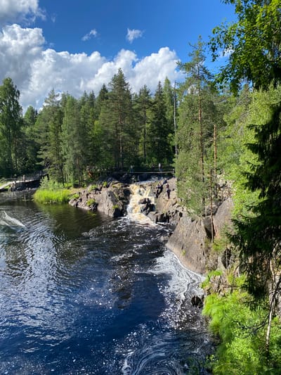The image depicts a picturesque natural scene featuring a waterfall cascading over rocks into a dark, turbulent body of water. The waterfall is situated in a rocky gorge surrounded by lush green vegetation and tall pine trees, characteristic of the Ruskeala Mountain Park in Russia. The sky above is a vibrant blue, dotted with fluffy white clouds, suggesting a sunny day.
Several people are visible in the image, some walking along wooden bridges that span across the gorge, while others are seen near the waterfall, possibly observing the natural beauty or taking a break. The water exhibits a frothy, swirling pattern near the edge of the rocks, indicating the force of the falling water. The overall impression is one of a serene and attractive natural landscape, popular for outdoor recreation.