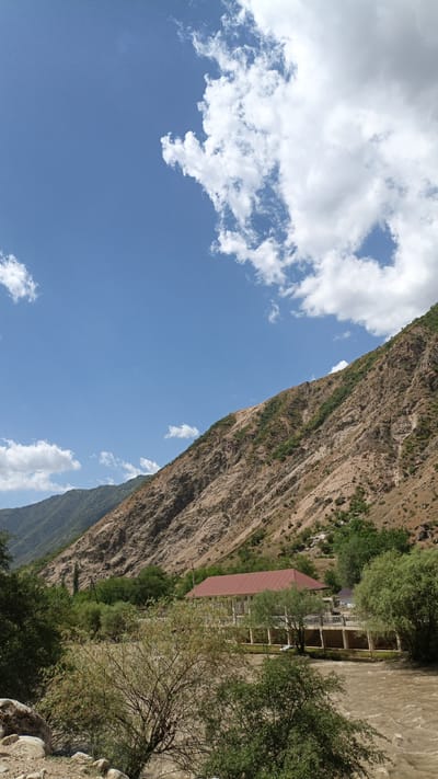 The image depicts a scenic mountain landscape with a river flowing in the foreground. A large, rugged mountain slope dominates the right side of the frame, characterized by exposed rock and sparse vegetation. The slope is angled steeply upwards, with a clear division between the arid rocky terrain and the greener, more vegetated foothills on the left. Above the mountains, a bright blue sky is filled with scattered white clouds, some of which are large and cumulus.

In the middle ground, a building with a red roof is visible, situated near the riverbank. The building appears to be a house or a guesthouse, with a veranda or balcony surrounding it, supported by pillars. A fence runs along the property line. Various trees and bushes are scattered throughout the scene, particularly along the river and in the area surrounding the building. The river itself is brown and appears to be flowing with some force, indicated by the turbulent water surface. The riverbed consists of rocks and pebbles. The overall environment suggests a rural or natural setting, likely in a mountainous region. While the prompt mentions Tavish, Tajikistan, the image itself does not contain any explicit geographical markers or features that definitively identify it as such.