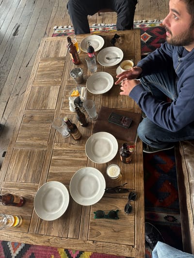 An overhead shot shows a group of people sitting at a wooden table in Stepantsminda, Georgia. The table is set with plates, glasses, and bottles of beer. A man in a blue hoodie and jeans is visible, holding a phone. Another person's legs are visible in the upper left corner of the frame, wearing black pants. The table has a rustic finish, with visible wood grain and some signs of wear. The overall atmosphere suggests a casual gathering or meal.