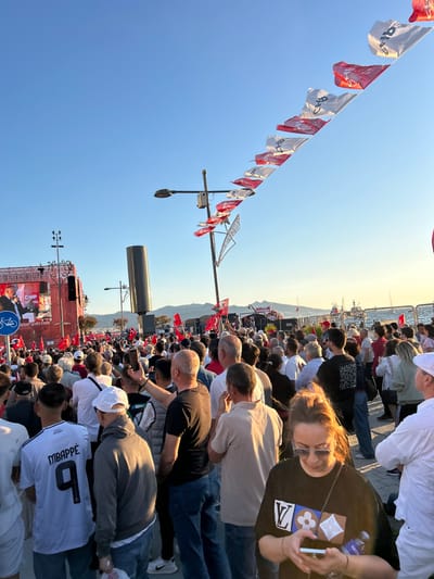 The scene is a crowded public square in İzmir, Türkiye (latitude: 38.437, longitude: 27.141), likely Konak Square. A large crowd of people are gathered, many of whom are holding red and white flags. The flags are flying in the breeze, while a speaker is addressing the crowd from a stage in the background. The people are listening intently, some holding phones and others making small talk with people nearby. There are some trees and a large building behind the crowd. The sky is blue with a few fluffy white clouds. It is a bright, sunny day.