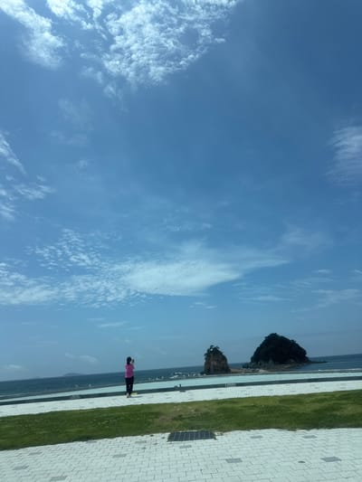 In Anmyeon-eup, South Korea, a person stands on a paved walkway facing the ocean, holding a phone to take a photo. The person is wearing a pink top and dark pants. In the background, the ocean stretches out to the horizon, with two small, rocky islands covered in vegetation visible in the water. A bright blue sky with scattered white clouds fills the upper portion of the image. The foreground consists of a paved area with a grassy strip beyond it.