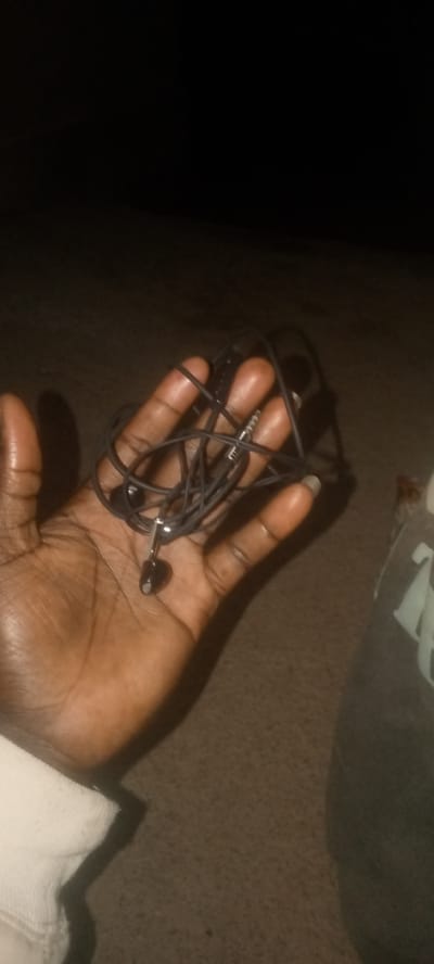 A dark-skinned hand with a white sleeve is holding a tangled set of black earbuds. The earbuds have a standard 3.5mm audio jack. The background is dark and appears to be a rough surface, possibly the ground or a carpet. The image is underexposed, with most of the scene obscured by shadow. The location in Zot Shen, Nigeria is not discernible from the image.