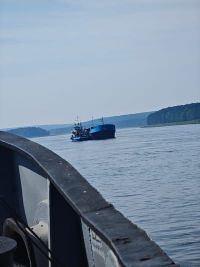 Two blue vessels are on the water. The larger vessel is a barge, and a smaller tugboat is attached to its stern. The vessels are in the middle of a wide body of water, with a treed shoreline in the background. The sky is clear and blue. In the foreground, a dark metal railing is visible, suggesting the image is taken from another vessel. This scene likely depicts activity on the Svir River in Podporozh'e, Russia, a city known for its river port and connections to the White Sea-Baltic Canal.