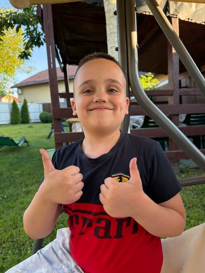 A boy, appearing to be around 8-10 years old, is shown from the chest up, seated outdoors in what seems to be a backyard. He has short brown hair, light skin, and green eyes. He is smiling with a dimple visible on his left cheek. He is wearing a t-shirt that is black on the top half and red on the bottom half, with a black "Ferrari" logo on the upper chest. His hands are raised, and he is giving a thumbs-up with both hands.
The background suggests a residential area, possibly Uhornyky, Ukraine, given the presence of a house with a red-tiled roof and green fencing. There is lush green grass, trees, and a wooden gazebo or structure behind the boy. A metal swing set is partially visible to his right. The lighting indicates it is daytime, with sunlight casting shadows.