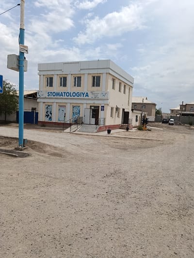 The image shows a two-story building with the sign "STOMATOLOGIYA" on the upper level, indicating it is a dental clinic. The building is light beige with white trim and multiple windows. In front of the building, there is a paved entrance with stairs and a railing. Three posters are visible on the lower level, two of which appear to show images of smiling people with healthy teeth. A blue lamppost with a white sign is located to the left of the building. The scene appears to be in a semi-urban or rural setting, with unpaved ground and a few other buildings in the background. The sky is partly cloudy. The location is identified as the city of Kungrad, Uzbekistan. There are no people visible in the image.