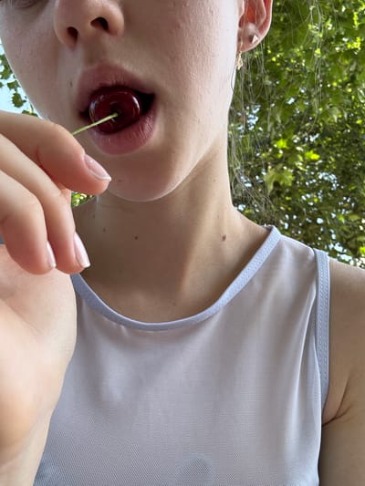 This is a close-up, low-angle shot of a person's mouth and shoulder area, with a cherry held by their fingers and partially inserted into their mouth. The person has fair skin and is wearing a light-colored, sheer top. Their lips are slightly parted, revealing the dark red cherry. Their fingernails are neatly manicured and painted with a light, neutral color. In the background, out-of-focus green leaves suggest an outdoor setting with dappled sunlight, hinting at a warm, possibly summer day. The overall mood is playful and sensual, with a focus on the cherry and the act of holding it in the mouth. The image appears to be taken in Bürgstadt, Germany, likely outdoors during daylight hours. There is no visible text in the image.