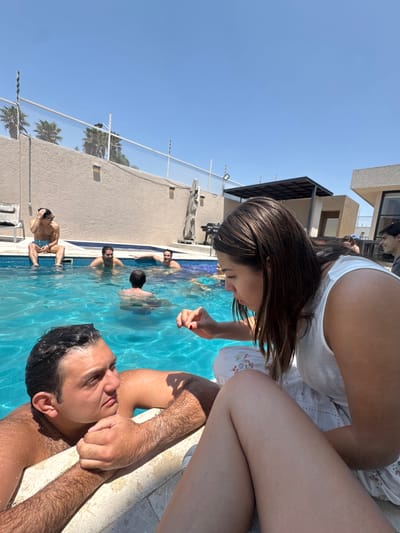 The image shows a group of people at a swimming pool on a sunny day, likely in Santa Cruz Tehuixpango, Mexico. A man is lying on the edge of the pool, partially submerged, with his head near the camera. A woman is sitting on the pool's edge, leaning over him. Other people are swimming and standing in the pool. The pool is surrounded by a wall with a wire fence and some buildings. The sky is clear and blue.