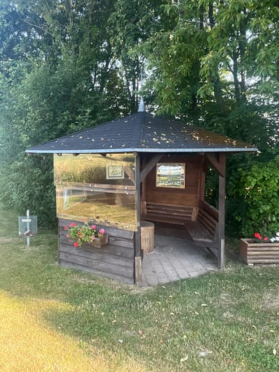 The image shows a small wooden hut with a dark shingled roof in a garden setting. The hut appears to be an outdoor shelter or playhouse. It has wooden walls and a transparent front panel, possibly made of plexiglass, which reflects the surrounding greenery. Inside the hut, a small wooden picture frame is visible.
Attached to the side of the hut is a wooden bench with slats, and in front of it, a wooden structure with a log-like element. A small planter with pink flowers is attached to this wooden structure. To the right of the hut, a paved walkway is visible, leading away from it. Beyond the walkway, a grassy area with scattered leaves is seen. The background is dominated by lush green trees and foliage, suggesting a natural outdoor environment.
A metal bin with a lid, possibly a trash can or a receptacle for dog waste, is positioned on the grass in front of the hut. The overall impression is one of a quaint, perhaps child-friendly, structure in a serene garden. The lighting suggests it is daytime, likely late afternoon due to the warm, golden hue on the grass. There are no people visible in the image. No visible text is present.