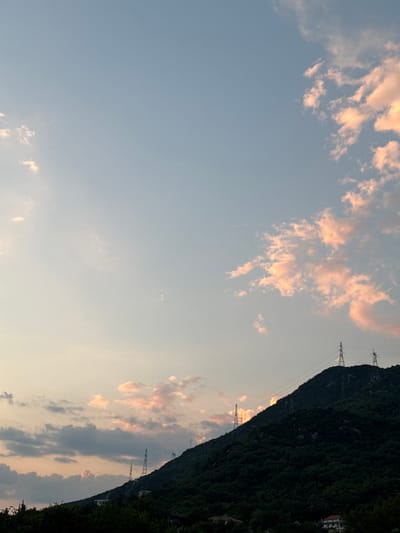 The image depicts a cloudy sky at sunset, with the sun's light casting a warm, pinkish glow on the clouds. The sky is a gradient of soft blues and oranges, suggesting the end of the day. A dense, dark green mountainside dominates the right side of the frame, its texture suggesting a heavily forested slope. Along the crest of the mountain, several tall, metal electricity pylons are visible, their stark geometric forms contrasting with the organic shapes of the mountain and clouds. These pylons are strung with power lines, indicating infrastructure present in the area.
Towards the top right of the image, a few buildings with white facades and red roofs are partially visible, nestled into the upper slopes of the mountain, suggesting a small settlement. The overall scene evokes a sense of natural beauty at dusk, with human-made elements of power transmission integrated into the landscape. The image appears to be taken from a low vantage point, looking upwards towards the sky and the imposing mountain. The weather is calm, with the clouds suggesting a tranquil end to the day. There is no visible text in the image.