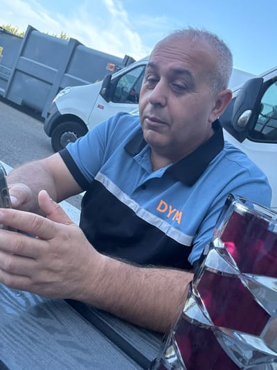 In Boom, Belgium, a middle-aged man with graying hair and olive skin sits at a table, looking down at his smartphone. He is wearing a blue polo shirt with a black collar and a horizontal black and white stripe across the chest, with the orange letters "DYM" visible on the blue portion. His hands are resting on the table as he holds the phone. To his right, a faceted glass object filled with a dark red liquid is partially visible. In the background, a grey dumpster is positioned next to a white van. The scene appears to be outdoors, possibly at a business or industrial location, with a clear sky overhead.