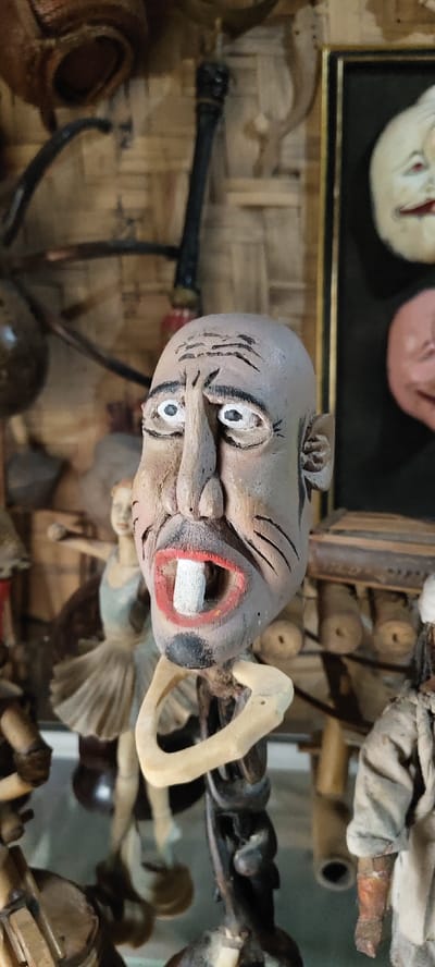 The image depicts a close-up, vertical shot of a collection of carved wooden figures and artifacts, located in Cikanyere, Indonesia. In the foreground and slightly right of center is a detailed wooden bust, portraying a bald head with a light brown-grey complexion. This figure features a prominent, long nose, wide-open eyes with white sclera and black pupils, distinct forehead wrinkles, and a wide-open mouth displaying red lips and a single, large, rectangular white tooth. Short black lines are carved on the cheeks, resembling sideburns or marks. The bust is affixed to a dark, intricately carved stand, with a light-colored, bone-like curved element encircling its neck.
In the blurred background to the left, a full-body wooden figurine resembling a ballerina in a pale tutu is visible, posed mid-dance. To the right, also blurred, is a dark-framed artwork containing at least two relief masks or faces, one white and one pinkish, with distinct features. The overall background consists of a woven light brown material, consistent with bamboo or rattan matting, and several other partially visible, out-of-focus carved wooden objects and tools are scattered throughout the scene. Part of another fabric-dressed figure is visible in the lower right corner. The scene suggests an indoor display or workshop environment for traditional crafts.