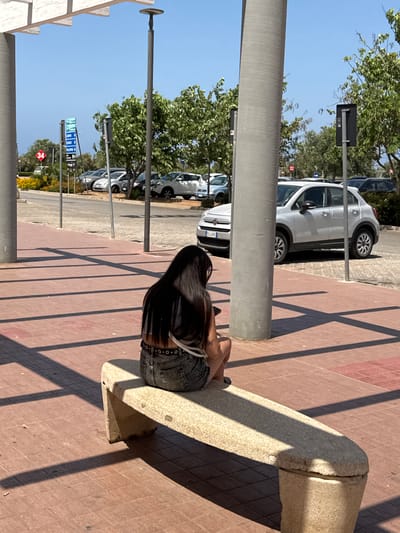 A person with long black hair is sitting on a stone bench outdoors. They are wearing denim shorts and a light-colored top. The scene appears to be a parking lot with several cars visible, including a white hatchback. Trees and a clear blue sky suggest a sunny day. The ground is paved with reddish-brown tiles, and long shadows are cast by overhead structures, indicating it is likely midday. Traffic signs are visible in the background, one with the number "34" in a red diamond shape. Another sign has arrows and text that appears to be in Italian. The setting is likely a public area, possibly near a commercial establishment or a road. The person's posture suggests they are relaxed, perhaps taking a break.