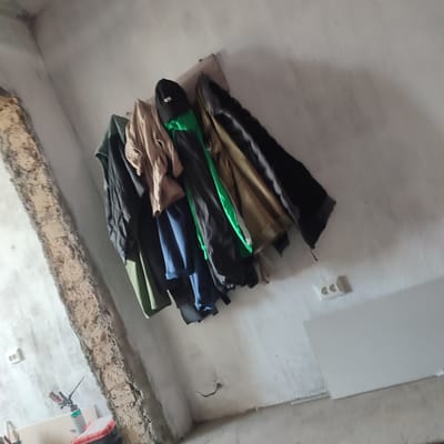 A somewhat blurry, low-angle photograph captures a wall in what appears to be a construction or renovation site in Alga, Kyrgyzstan. A rough, unfinished concrete column or beam runs diagonally from the lower left to the upper left of the frame. A rack on the wall to the right holds several items of clothing, including dark jackets, a tan shirt, and a bright green item, possibly a jacket or scarf. Two white electrical outlets are visible on the wall. In the lower left, a red and black tool with a spray nozzle is resting on what appears to be a red and black container. The lighting is dim, and the overall impression is one of disarray or unfinished work.
