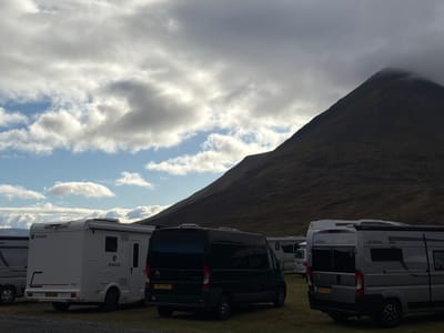 The image shows a parking area in the Isle of Skye, United Kingdom, filled with campervans. The background is dominated by a large mountain with its peak partially obscured by clouds. The sky is overcast with patches of blue. Several campervans of varying sizes and colors are parked in the foreground. From left to right, the campers include one white Rimor Sarus with license plate BYX26 AUE, a black Citroen Relay van with license plate CK17 GVC, and a gray Pössl Summit van with license plate HST-37-H. Other vans are visible further back in the parking area. The ground is covered with short vegetation.