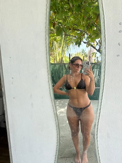 This image is a mirror selfie taken by a woman in a tropical outdoor setting. The woman is reflected in an organically shaped mirror, which has a textured white frame adorned with small shells or pebbles along its top edge. She is wearing a dark two-piece bikini with a golden shell-shaped accent on the top. Her hair is pulled back, and she is wearing dark-rimmed sunglasses. She holds a light-colored smartphone with her right hand to capture the photo.

The reflection in the mirror reveals a bright, sunny day with a clear sky and abundant lush green tropical foliage, including what appear to be palm trees and other large-leafed plants. The woman is standing on a light-colored surface, possibly sand or light-colored pavement, which is also reflected in the mirror.

The activity taking place is a self-portrait (selfie). The time of day appears to be daytime, given the bright illumination. The weather is sunny and clear, indicative of good conditions for an outdoor shot in a tropical location like Thoddoo, Maldives. No visible text can be discerned in the image.