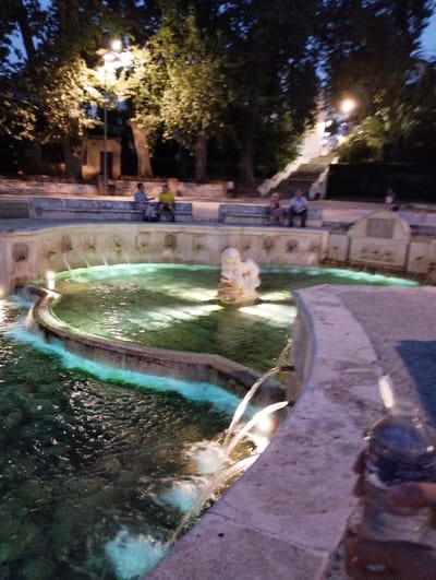 This is a nighttime photograph of the Fuente del Rey in Priego de Córdoba, Spain. The fountain is centrally located, with water cascading from numerous spouts into a large, circular pool. The water is illuminated by green lights, creating a vibrant glow. Several people are visible in the background, sitting on benches and walking near a staircase that ascends a gentle slope. Trees surround the fountain, their leaves dark against the twilight sky. The overall scene is one of peaceful, illuminated public space at dusk.