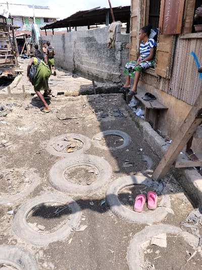 The scene depicts a narrow, unpaved alleyway within a densely populated area of Oworonshoki, Nigeria, under daylight conditions. The ground consists of uneven dirt, interspersed with numerous worn-out vehicle tires partially embedded, likely serving as stepping stones or ground stabilization. Various debris, including plastic bags and general refuse, is scattered along the path and in crevices.
On the right side, a building constructed from weathered wooden planks and other materials features an open window opening. A young woman is seated sideways within this opening, wearing a blue and white striped short-sleeved top and a patterned skirt. She has dark hair, possibly braided, and wears dark sandals on her feet. Her gaze is directed to the left side of the frame. Below her, a wooden bench holds a pair of black sandals and accumulated trash. A bare foot with an ankle chain is partially visible near the bottom right, next to a wooden support beam.
On the left side of the alley, a person, likely female, dressed in a green garment (possibly a wrapper) and carrying a black backpack, is walking away from the camera. Her feet appear to be wearing light-colored open-toed sandals or are bare. Further down the path, several other individuals, some also in green attire or carrying bags, are visible walking in the same direction.
In the mid-ground, separating the walking path from the right-side building, there is a low concrete block wall, exhibiting wear and discoloration. The background reveals additional residential or commercial structures with corrugated metal roofs. One roof in the far background appears to have solar panels installed. The overall environment suggests an informal settlement or community area characterized by rudimentary construction and visible refuse.