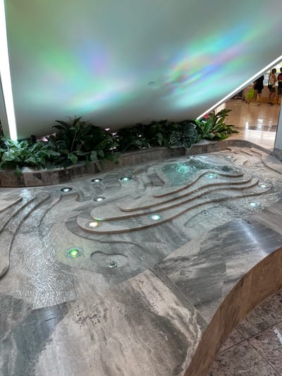 The image shows a water feature in a public space located at coordinates -27.5, 152.973 in Indooroopilly, Australia. The water feature is made of grey stone and consists of a series of curved channels and pools with water flowing through them. The water appears to be illuminated from below by a series of small, round, white lights. There is also a larger circular white light on the ceiling of the space above the water feature. The water feature is located in a semi-enclosed space with a low wall, and there are potted plants on the wall. The space is tiled in grey tiles and the floor slopes down towards the water feature. The space appears to be a shopping centre or public mall. There are no people in the image.