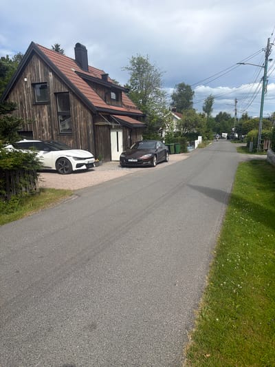 A residential street in Lørenskog, Norway, is depicted under a cloudy sky. In the foreground, a paved road runs diagonally into the distance. To the left of the road, a modern wooden house with a red tiled roof is partially visible, with two cars parked in its driveway: a white electric vehicle and a dark-colored sedan. The white car has license plate "ED 37165". The dark sedan has license plate "EL 63110". Green refuse bins are situated on the side of the road. Further down the street, a person is riding a bicycle, casting a shadow on the road. The street is lined with trees and houses on both sides, with power lines and a lamppost visible. Lush green grass borders the right side of the road.