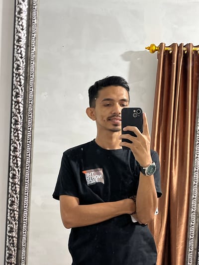 A male individual is depicted in a mirror selfie taken indoors in Alueduamuka S, Indonesia. He possesses dark, neatly styled hair, dark eyes, a slight smile revealing upper teeth, a small mustache, and a goatee. He is wearing a black short-sleeved t-shirt with a reversed "THE NORTH FACE" logo visible on the left chest. A black smartwatch with a round face and green accents is secured on his left wrist, and multiple dark-colored beaded bracelets are worn on his right wrist. He holds a black smartphone, featuring a multi-lens camera array, in his right hand, positioned to capture his reflection. His left arm is crossed over his chest. The mirror features an ornate, possibly silver-colored or distressed metallic, carved frame with scrollwork designs along its left edge. The background consists of a light-colored, textured wall. Brown, pleated curtains hang from a golden rod with a decorative finial, visible on the right side of the frame. The scene is illuminated by an unseen indoor light source.