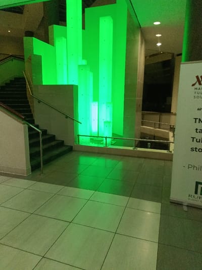 The image shows an indoor space illuminated with green lighting. A staircase ascends alongside a decorative arrangement of vertical light panels. Light reflects on the tile floor. In the background, a multi-level structure with metal railings is visible through an opening. A banner with logos and text occupies the right foreground.