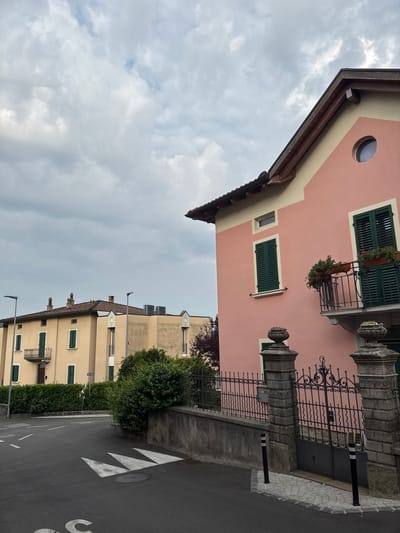 In the city of Balerna, Switzerland, on a cloudy day, a street corner is depicted with two buildings. The building in the foreground is a two-story structure painted a light pink with cream trim. It features dark green shutters on its windows, one of which is a round porthole window near the roofline. A small balcony with a wrought iron railing and planters adorns the second story. The building is bordered by a stone wall with decorative urns and a black wrought iron gate. The building in the background is a lighter yellow color and also features dark green shutters and balconies. A dense green hedge lines the street in front of this building. The street itself is asphalt with white road markings, including a crosswalk and what appear to be speed reduction markings. The sky is overcast with various shades of gray and white clouds.