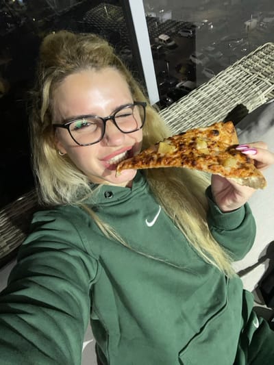 A young adult female, with long blonde hair styled partially in an updo, wearing black-rimmed eyeglasses and a dark green Nike hooded sweatshirt, is captured in a selfie. She is biting into a triangular slice of pizza topped with cheese and pineapple. Her left eye is closed in a wink, while her right eye is open, directed towards the camera. Her right hand, featuring pink and white manicured nails, holds the pizza slice. A small silver nose piercing and a silver hoop earring in her visible ear are present. She is seated on or beside a light-colored, woven outdoor chair or sofa. The background displays a dark, nighttime urban landscape, suggesting an elevated position such as a balcony, overlooking a parking lot with discernible vehicle lights in the city of Yavne, Israel. A white vertical architectural element is visible on the left side of the frame.