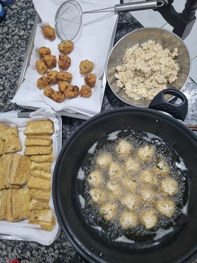 This media file captures a culinary scene, likely in a home kitchen in Laranjal Paulista, Brazil. The primary subjects are various food items being prepared for frying.

In a metal bowl, a mixture of crumbled food, possibly a batter or filling, is visible. Next to it, a black frying pan contains numerous small, spherical food items submerged in bubbling hot oil, actively frying. To the left of the pan, on a white paper towel-lined tray, a collection of golden-brown fried fritters has been placed, accompanied by a metal skimmer. Below these fritters, another tray holds sliced, pre-fried tofu, also resting on paper towels.

The scene depicts the process of deep-frying, with the bubbling oil and the golden-brown hue of the fried foods indicating recent activity. The overall setting appears to be a countertop with a speckled granite-like surface. The presence of a wooden chair leg in the upper left corner suggests a domestic environment. There is no visible text in the image. The time of day and weather are not discernible from the image. The activity is purely focused on food preparation, without any human subjects present.