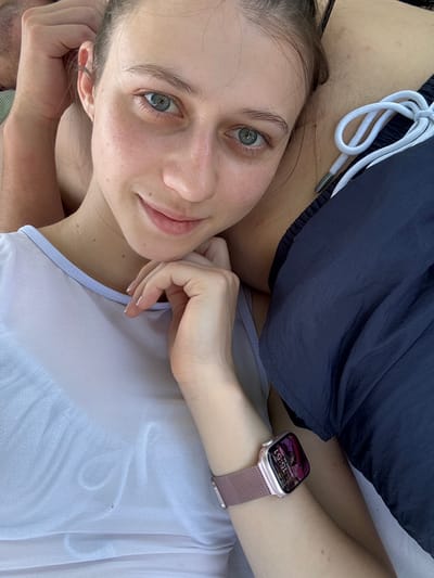 A young woman is lying down, resting her head on someone's shoulder and arm. She is looking at the camera with a slight smile. Her left hand is resting on her chin, and she is wearing a light-colored, sheer tank top. On her left wrist, she is wearing a rose gold Apple Watch, displaying the time as 3:51 PM and the date as Sunday, June 28. To her right, she is wearing navy blue athletic shorts with a white drawstring. The background is out of focus, but suggests an outdoor setting, possibly a park or beach. There is no discernible information within the image that places the scene in Bürgstadt, Germany.