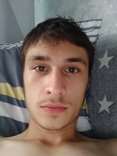 A close-up selfie, taken from a high angle, shows a young Caucasian male lying down on a bed. The man has brown hair, brown eyes, and light facial hair. He is wearing a light-colored t-shirt. The background consists of pillows and a wall. One pillow has blue and white stripes, and another is grey with white stars. The wall appears to be textured and light blue. The lighting suggests an indoor environment, possibly a bedroom. The image was likely taken in Мамлют қаласының әкімшілігі, Kazakhstan.