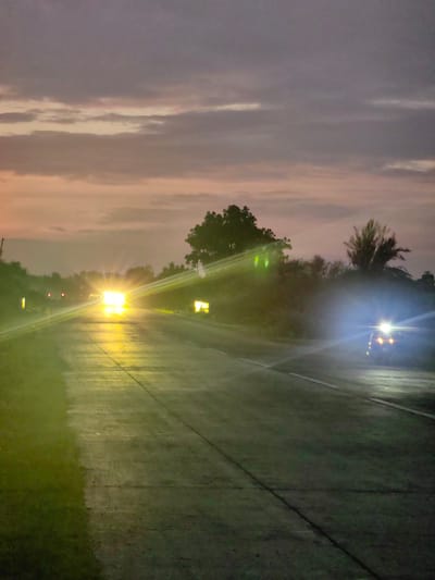 The image captures a twilight scene on a road, likely in or near Dattapur, India, given the context. A paved road stretches into the distance, reflecting the dim light of the sky and the headlights of approaching vehicles. The sky is a gradient of soft purples, pinks, and oranges, with scattered clouds. Dense foliage lines both sides of the road, with trees silhouetted against the colorful sky.
In the foreground, the road surface is dark and wet, with faint lines marking the lanes. On the left, a bright yellow light emanates from a source just off-camera, casting a strong reflection on the road. Further down the road, a vehicle with intensely bright headlights approaches, its light creating a powerful glare that washes out details. Another vehicle is visible on the right side of the road, also illuminated by bright headlights. The overall atmosphere is one of a transition from day to night, with the artificial lights of vehicles becoming prominent.