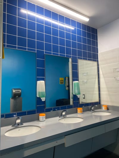 The image shows a public restroom in Virginia Beach, United States of America. The restroom features a row of three sinks with grey countertops and white porcelain basins. Each sink has a chrome faucet. On the countertops, there are two orange-lidded containers of what appears to be lotion or sanitizer. Behind the sinks, a wall is tiled with blue square tiles and white grout.
To the left, there is a bright blue panel with a silver hand dryer mounted on a grey cylindrical pedestal. Above the blue panel, a yellow and black sign with pictograms of a man and a woman is visible. Adjacent to this, a white soap dispenser is mounted on the blue panel.
In the center, a mirror reflects a blue door and a dark object on the floor. Another white soap dispenser is mounted on the blue panel to the right of the mirror. To the far right, a white tiled wall section is visible, with some clear plastic items mounted on it. The overall lighting is provided by fluorescent lights running along the ceiling.