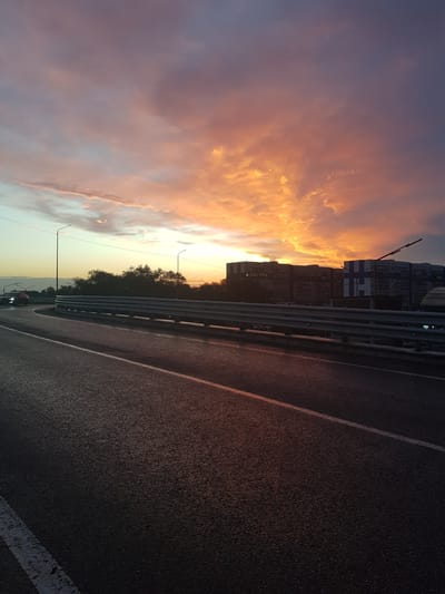 The image captures a road view in Бұлақты, Kazakhstan during sunset. The sky is ablaze with vibrant orange, pink, and purple hues, reflecting the dramatic cloud formations. In the foreground, a wet asphalt road with white lane markings curves to the left, suggesting a slight incline. A metal guardrail runs along the right side of the road. In the mid-ground, silhouetted trees form a dark line separating the road from a row of multi-story apartment buildings. Streetlights are visible on poles. A few cars are present on the road, with headlights on, indicating low visibility or it being twilight. The overall scene suggests a typical urban or suburban environment during a picturesque sunset.